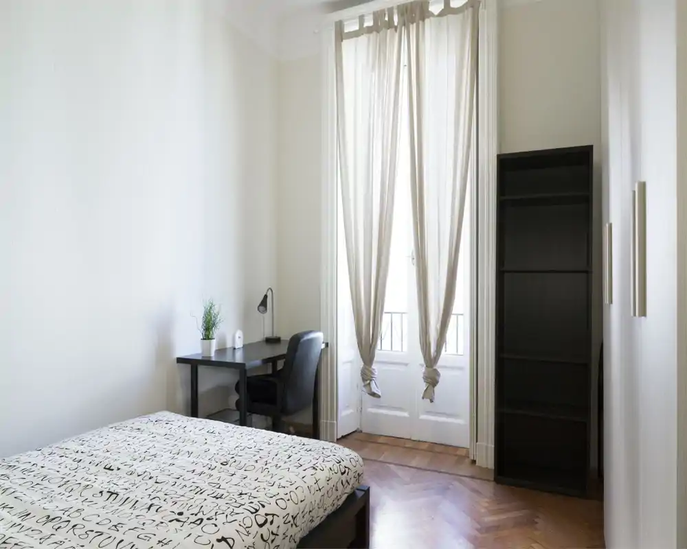 Chambre lumineuse et minimaliste avec un lit double confortable, un petit bureau et une chaise près d'une grande fenêtre laissant entrer une lumière naturelle douce.