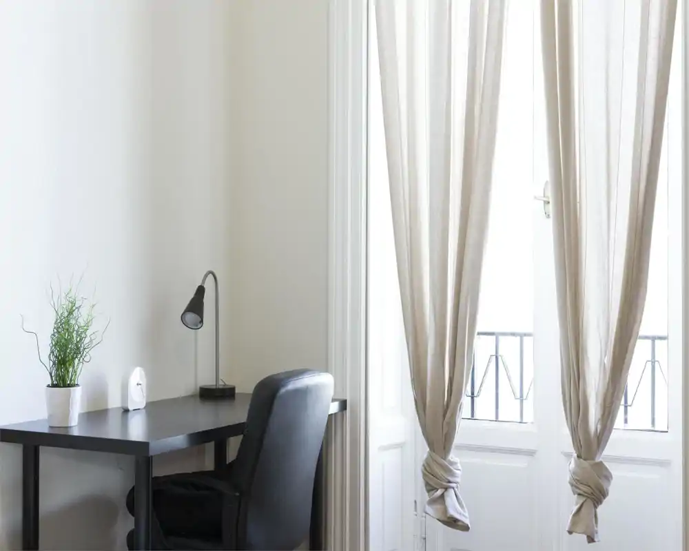 Coin salon lumineux et minimaliste avec un bureau noir, une lampe de bureau et un fauteuil confortable à côté de rideaux clairs et d'une porte-fenêtre.