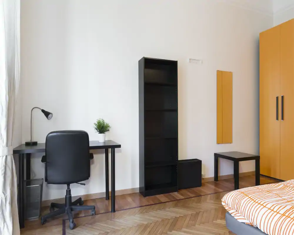 Dormitorio luminoso y minimalista con esquina de cama con ropa de cama a rayas naranjas, escritorio y silla negros y armarios a juego que ofrecen un espacio limpio y moderno.