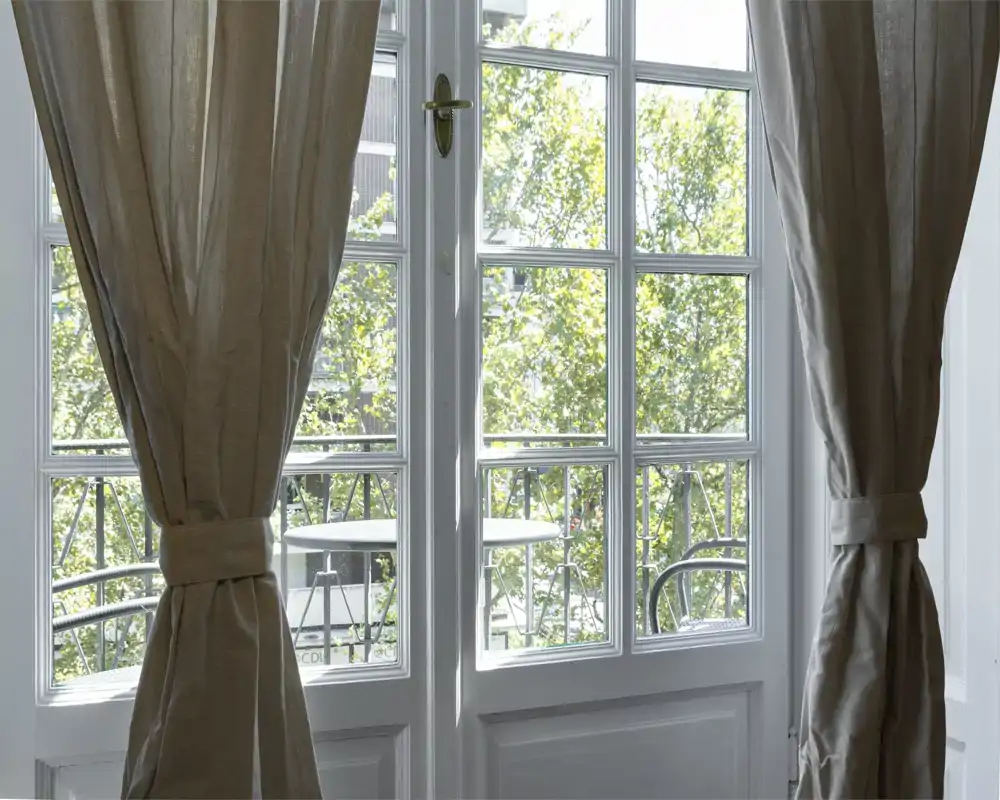 Balcón soleado visto a través de puertas francesas con cortinas recogidas, pequeña mesa redonda y sillas de metal visibles — un agradable espacio exterior comunicado con la habitación.
