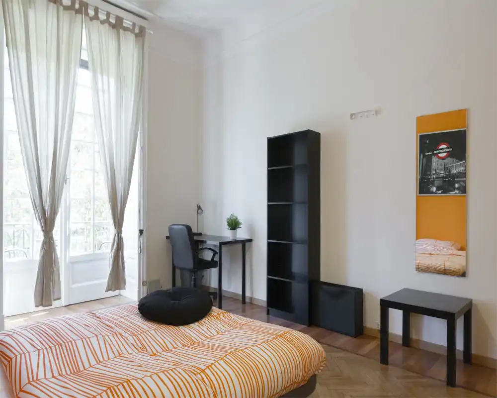 Dormitorio luminoso y moderno con cama doble con ropa de cama a rayas naranjas, escritorio y silla, ventana grande con cortinas translúcidas y estantería minimalista.