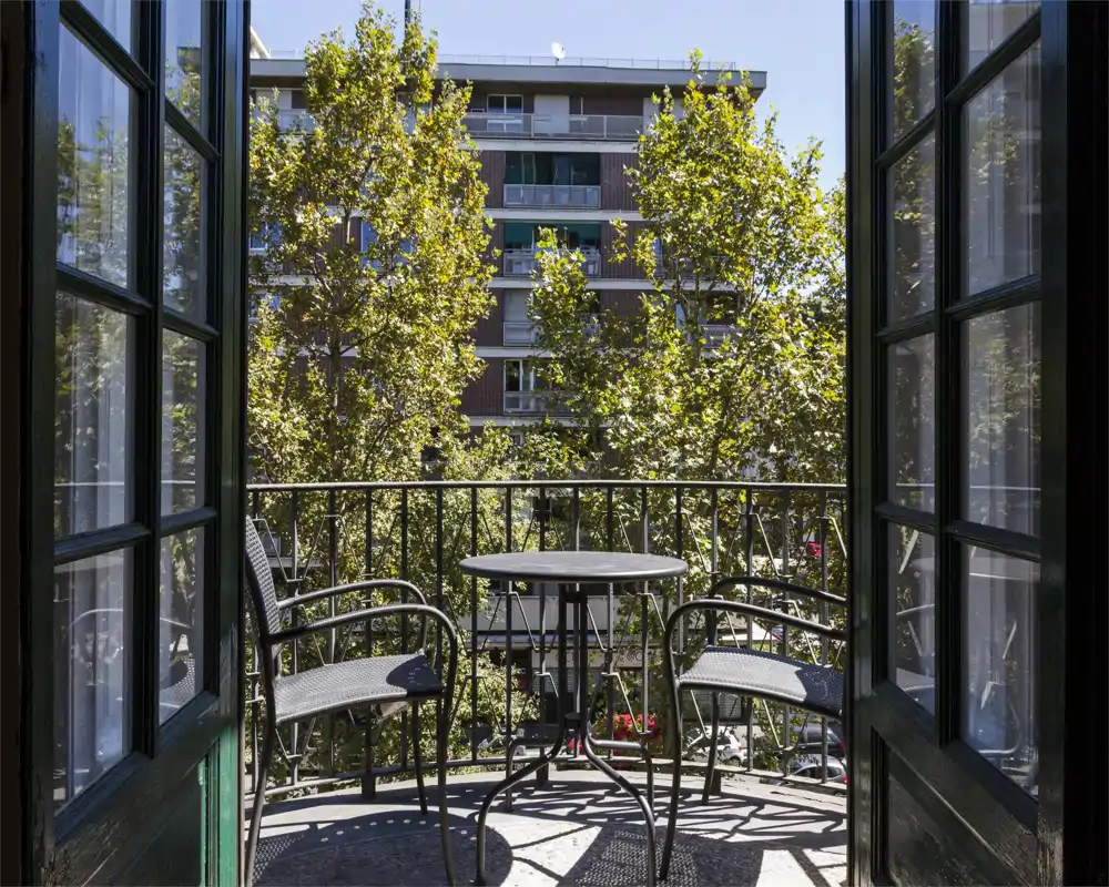 Balcón privado soleado con una pequeña mesa redonda y dos sillas, enmarcado por puertas ventanas abiertas y con vistas a árboles y edificios cercanos.