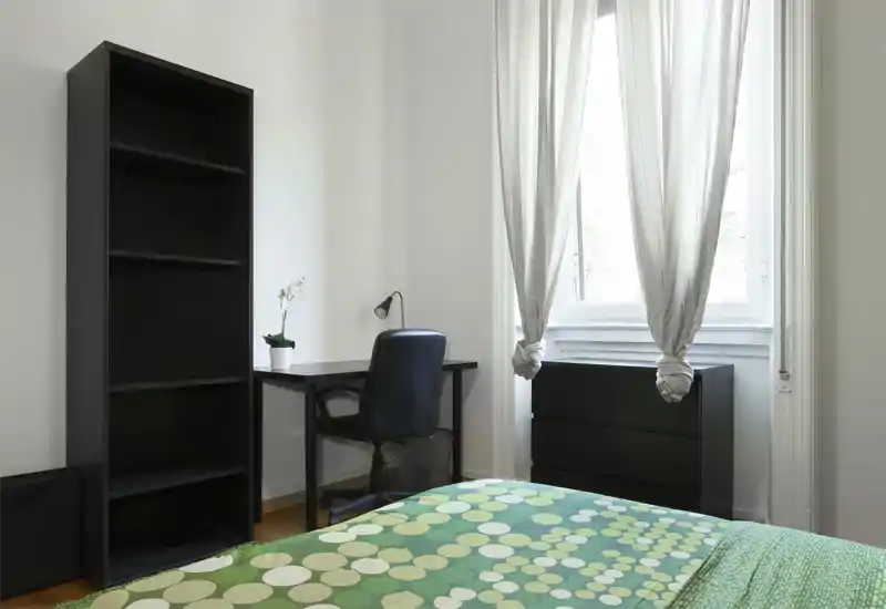 Dormitorio luminoso y minimalista con colcha verde estampada, un pequeño escritorio y silla junto a la ventana, y estantería y cómoda oscuras. La luz natural y las paredes neutras crean un rincón aireado para dormir y trabajar.