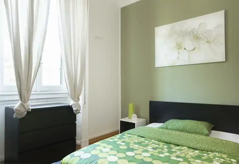 Dormitorio luminoso y bien presentado con cama doble con ropa de cama verde estampada, cabecero oscuro, mesita y luz natural procedente de una ventana grande.