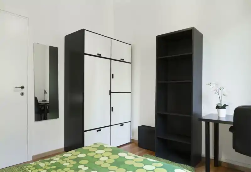 Dormitorio luminoso y minimalista con colcha verde estampada, armario alto blanco y negro, estantería y un pequeño escritorio con planta: limpio, moderno y bien iluminado.