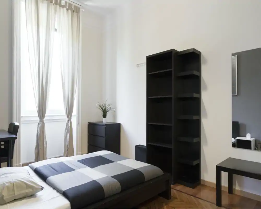 Chambre moderne et lumineuse avec un lit double couvert d'une couette à carreaux gris, grande fenêtre aux voilages légers et mobilier noir minimaliste (étagères, commode).