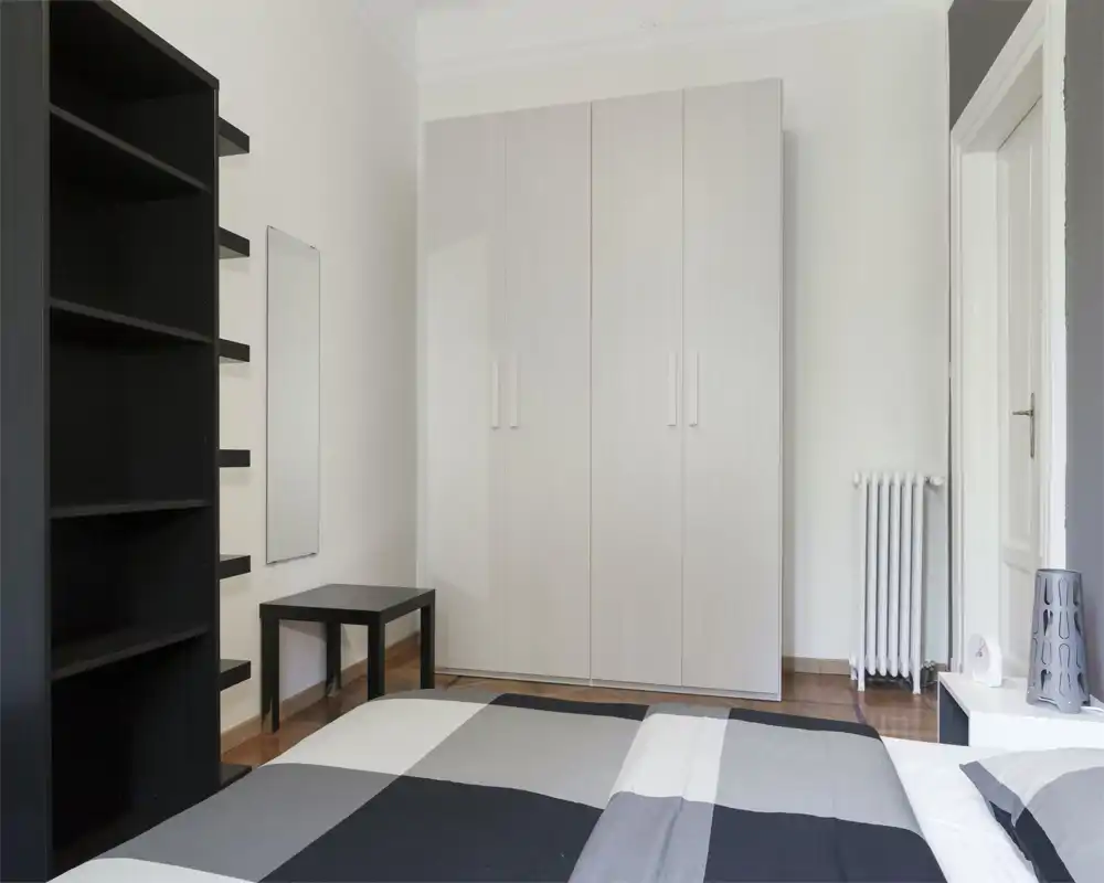 Chambre minimaliste avec lit double au premier plan, grande armoire blanche, étagères noires et mobilier sobre; tons neutres et lignes épurées pour un espace nuit moderne et apaisant.