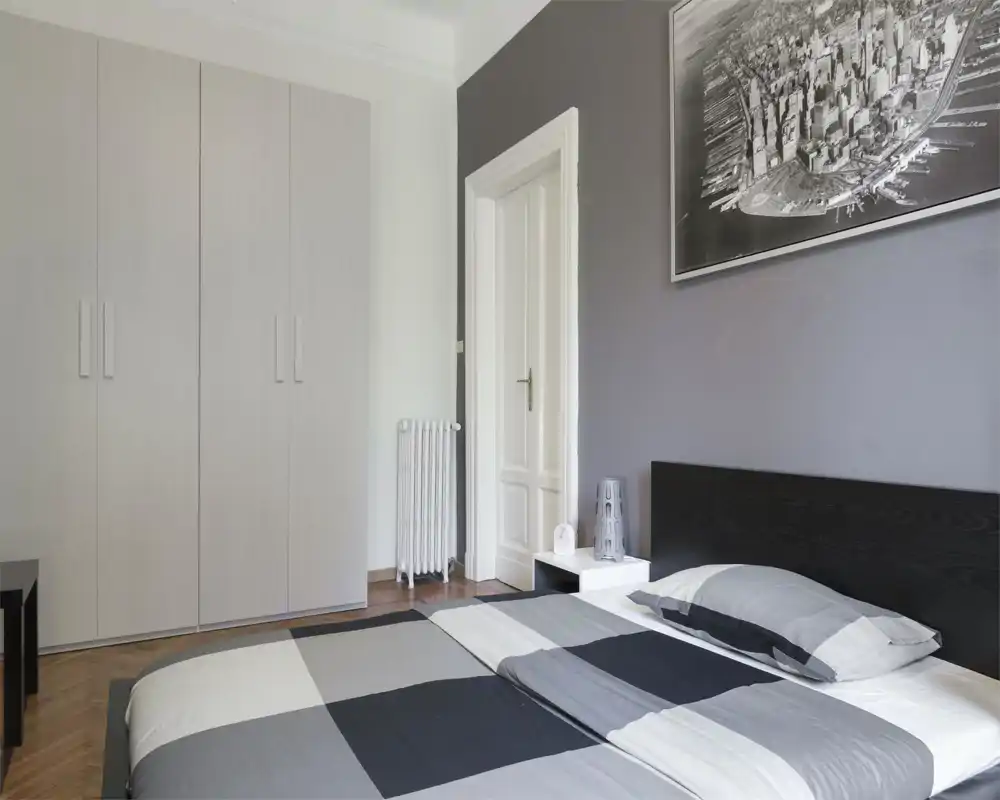 Chambre minimaliste avec grand lit en linge gris à motifs géométriques, tête de lit sombre, table de chevet avec lampe et armoire neutre — lumineuse, moderne et épurée.