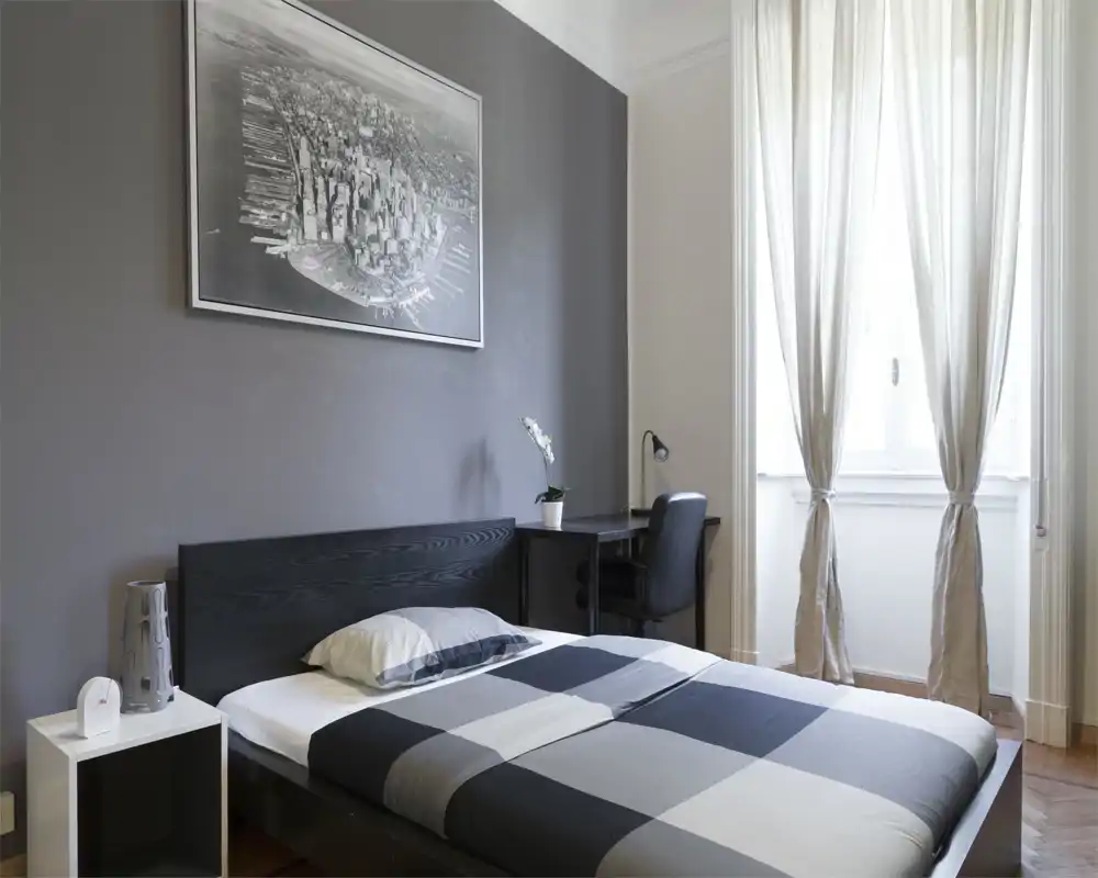 Chambre minimaliste et lumineuse avec lit double au linge de lit monochrome, tête de lit en bois foncé, bureau et chaise près d'une fenêtre claire avec rideaux légers.