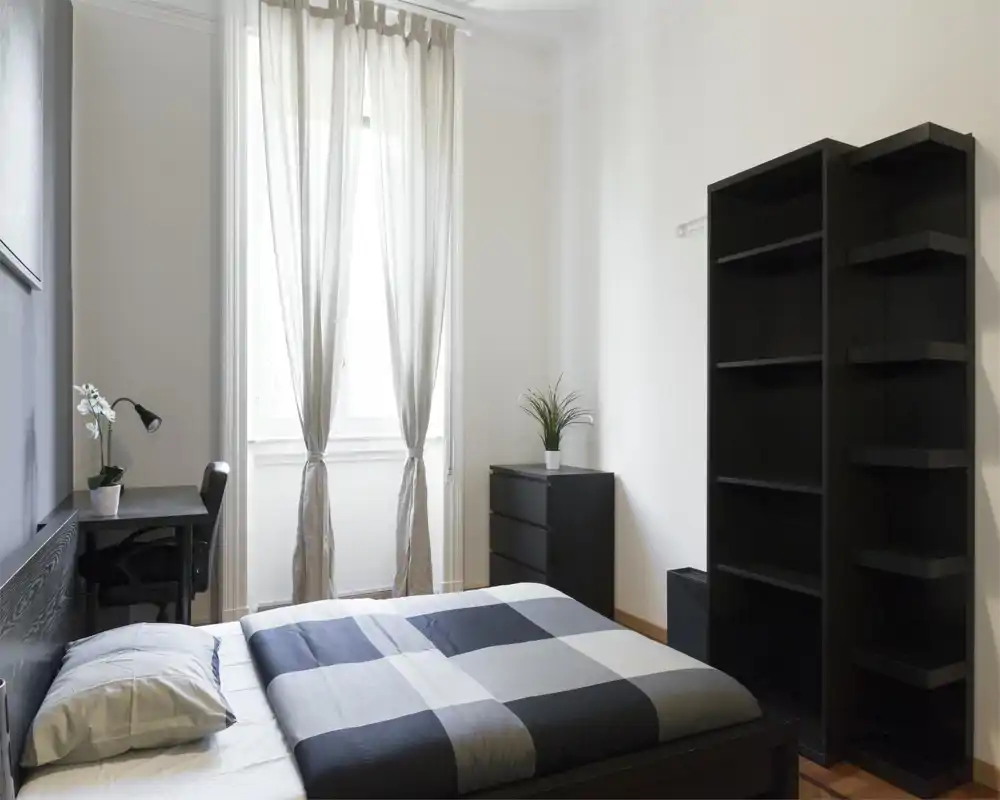 Chambre lumineuse et minimaliste avec un lit double au premier plan, mobilier noir sobre, commode et étagères, et une douce lumière naturelle provenant d'une grande fenêtre aux rideaux clairs.