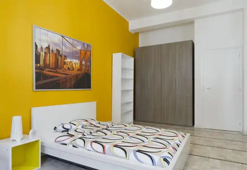 Bright modern bedroom with a low white platform bed, colorful patterned bedding and a striking yellow accent wall. Large wardrobe and shelving provide ample storage; framed city artwork completes the clean, contemporary look.