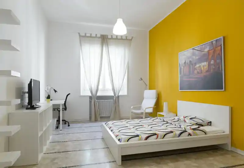 Bright, modern bedroom with a low white platform bed, patterned bedding, a comfortable armchair and a warm yellow accent wall that hosts framed artwork. The room includes a small desk and TV, plenty of natural light and clean minimalist styling ideal for rental listings.