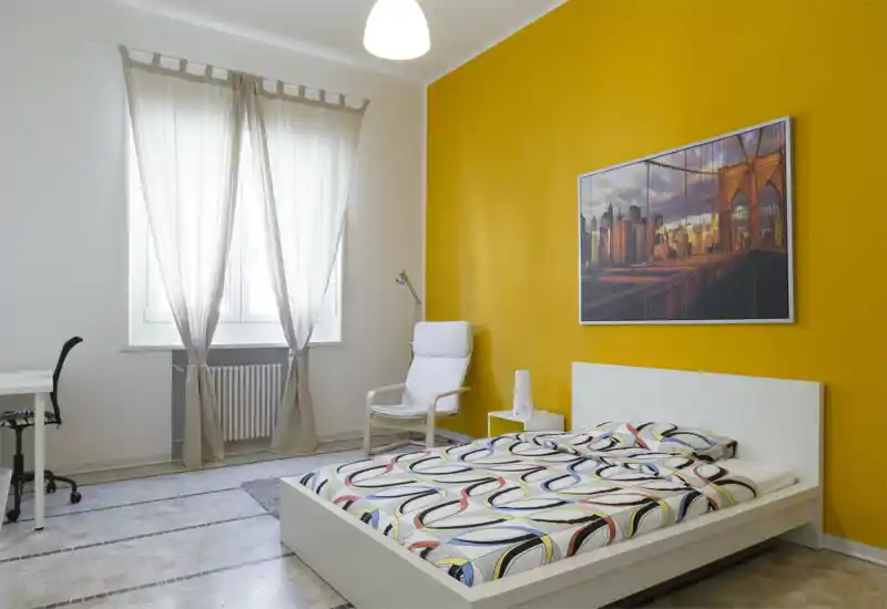 Bright, modern bedroom with a white platform bed, patterned bedding and a bold yellow accent wall. Natural light floods the room through sheer curtains; a comfortable armchair and small desk complete the space.