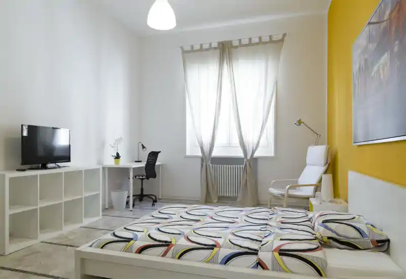 Bright, modern bedroom with patterned bedding, a comfortable armchair and a work desk by the window. The room features a yellow accent wall, large window with sheer curtains and neat minimalist storage.