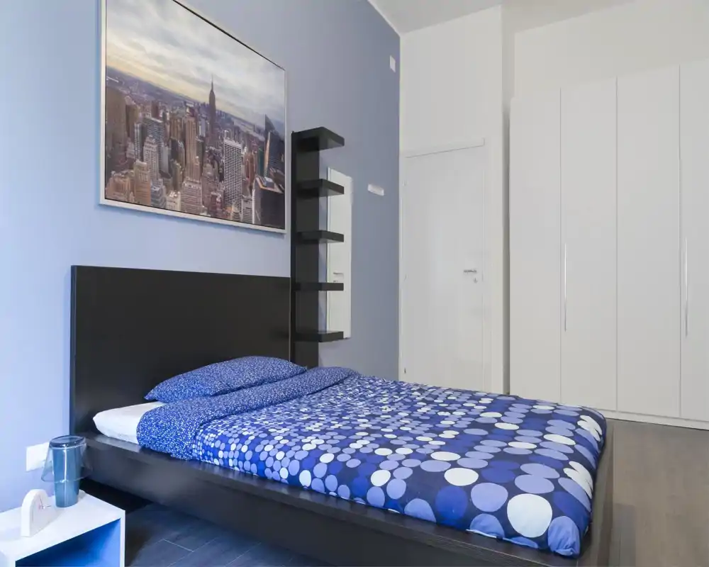 Camera da letto moderna e luminosa con un grande letto matrimoniale con copripiumino blu a fantasia, testata minimal e armadio bianco. La stanza ha una stampa con skyline cittadino e mensole semplici per uno stile contemporaneo e pulito.