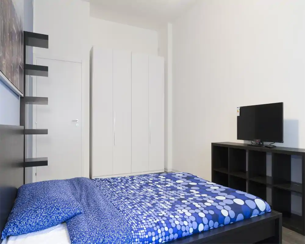 Camera da letto moderna e luminosa con letto matrimoniale con copriletto blu a motivo, armadio bianco e TV su mobile basso a scomparti — minimal, ordinata e ben illuminata.