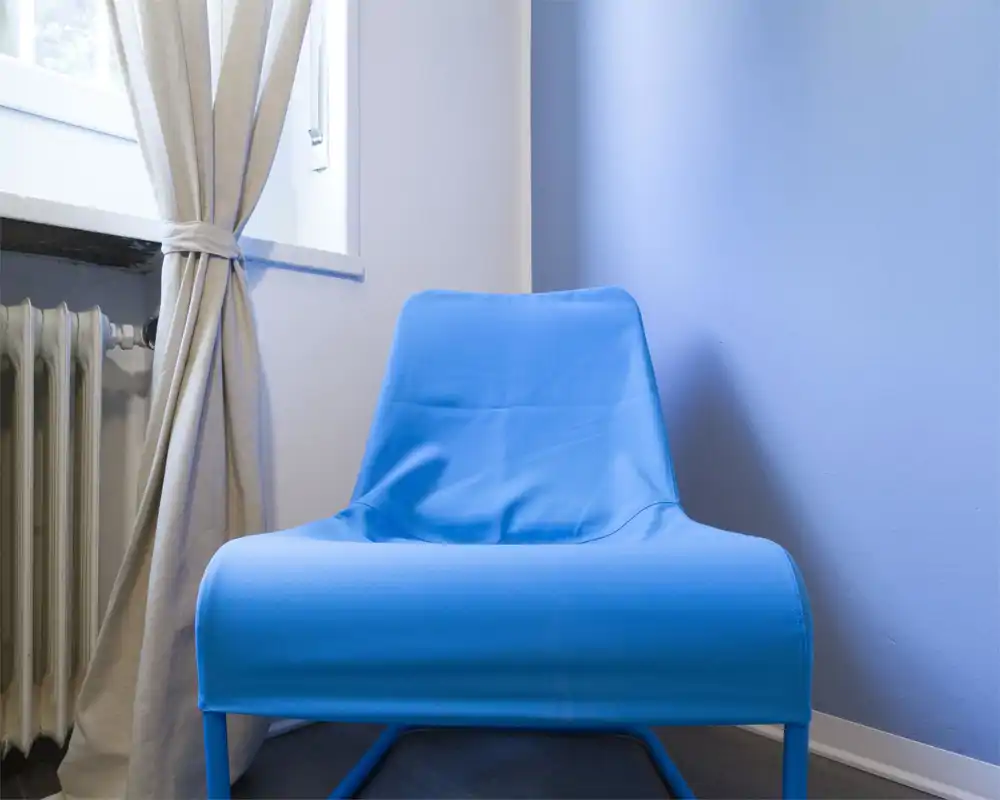 Angolo living minimal con poltrona blu brillante accanto alla finestra, tende neutre e termosifone — semplice, moderno e ben illuminato.