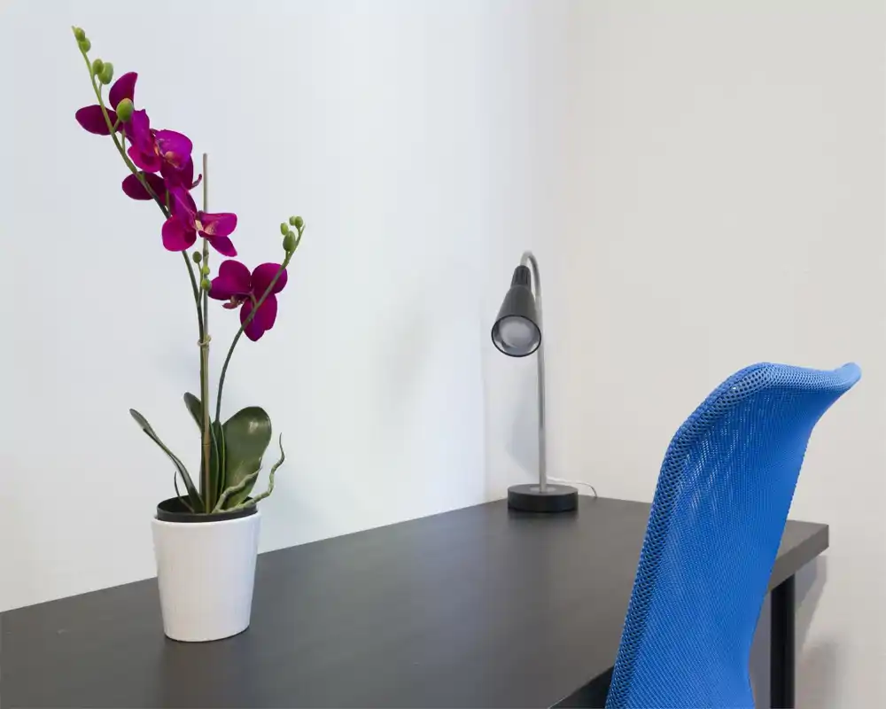 Angolo scrivania luminoso e minimal con sedia blu traforata, una lampada da tavolo e un'orchidea decorativa su un piano scuro — spazio di lavoro ordinato ideale per concentrazione.