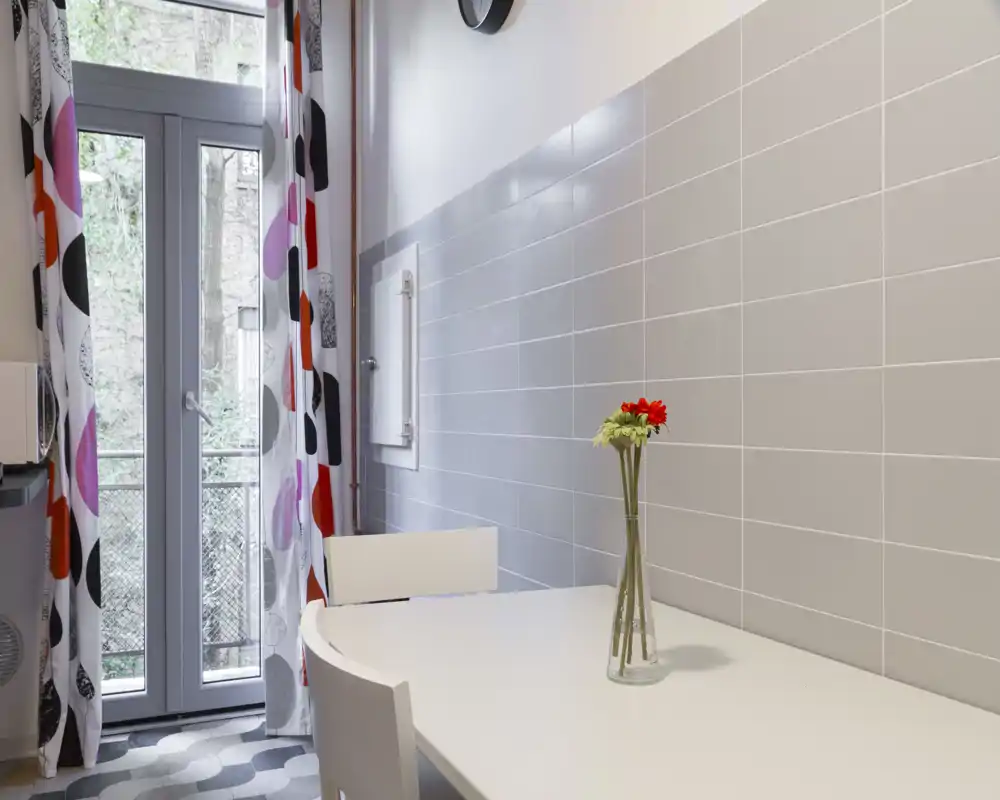 Angolo pranzo moderno e luminoso con tavolo bianco, sedie semplici e un piccolo vaso di fiori vicino a una porta finestra che apre sul balcone. Parete con piastrelle grigie e tende a fantasia per un aspetto pulito e contemporaneo.