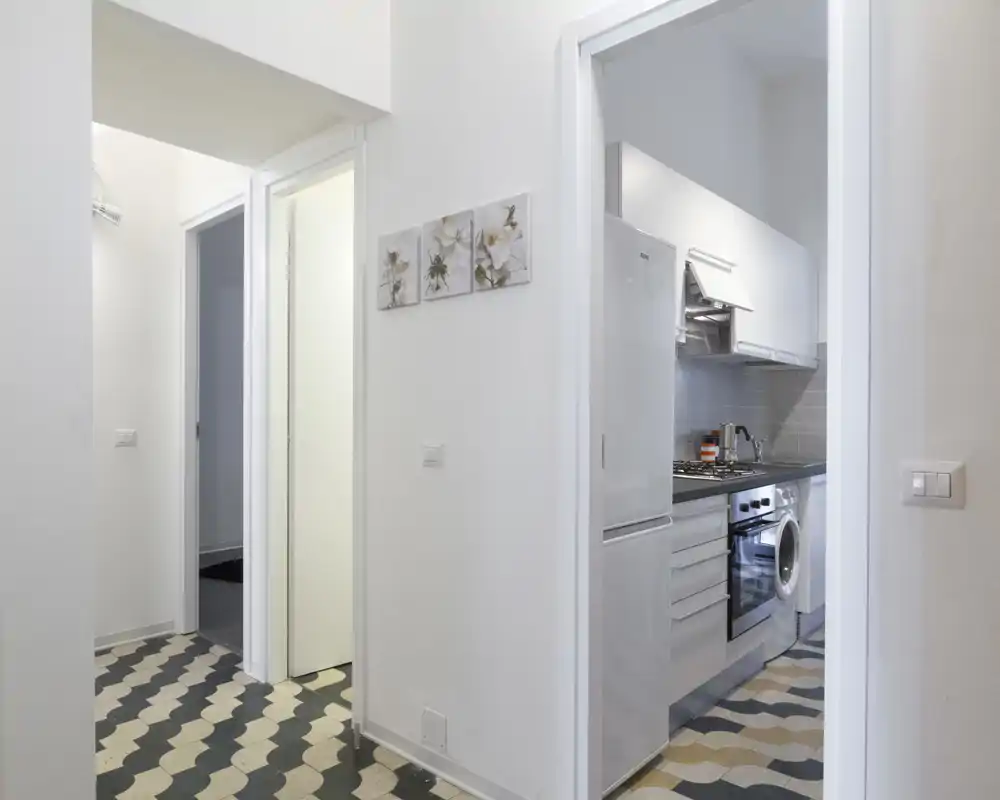 Cucina moderna e compatta visibile dalla porta, con elettrodomestici in acciaio, forno, lavatrice e pavimento piastrellato a motivo.
