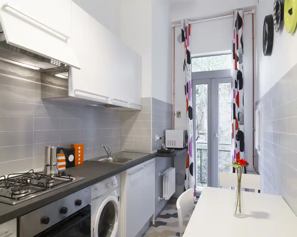 Cucina moderna e stretta con piano cottura incorporato, lavello, lavatrice e un piccolo tavolo da pranzo vicino a una porta vetrata che lascia entrare luce naturale.