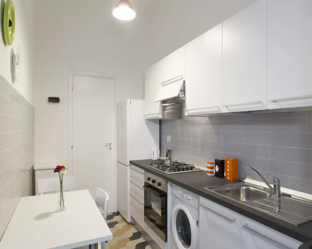 Cucina moderna e compatta con pensili bianchi, forno integrato, piano cottura a gas e lavello; ambiente luminoso e ordinato con un piccolo tavolo da pranzo e dettagli decorativi.