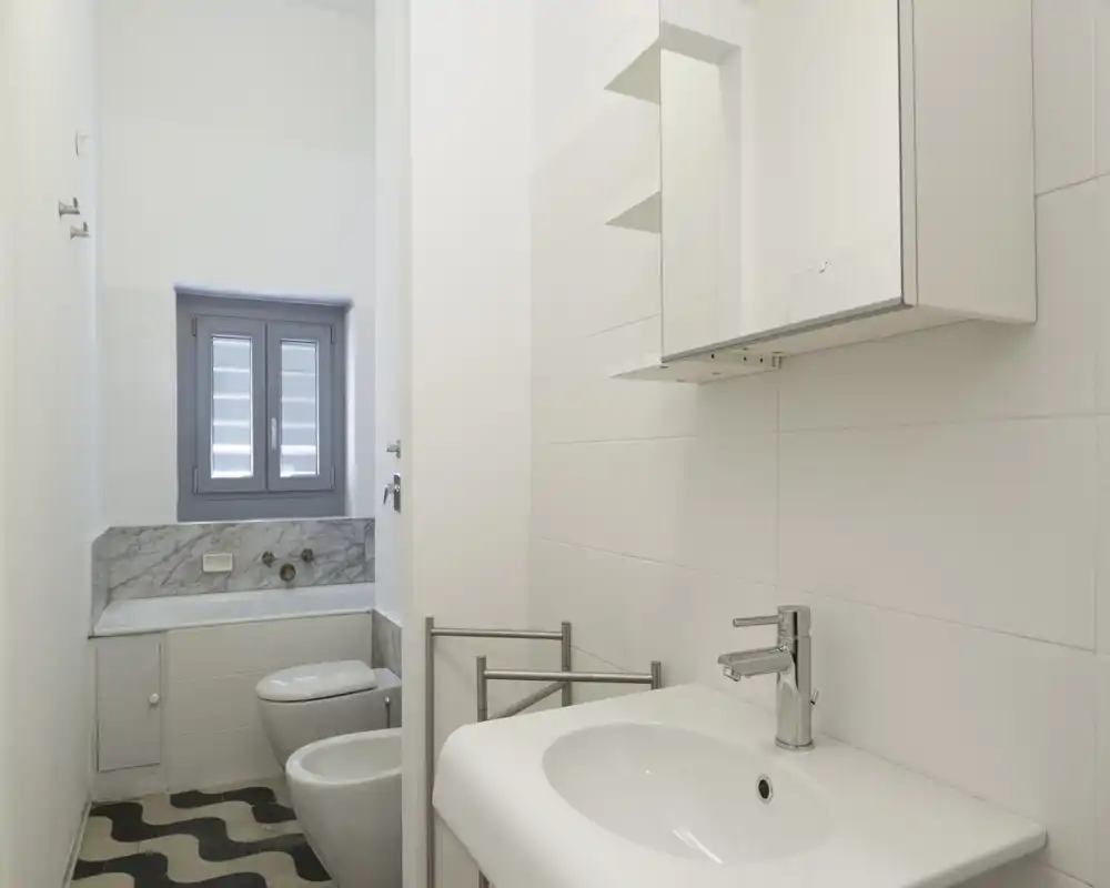 Bagno compatto e luminoso con lavabo a parete, bidet, wc e vasca sotto una finestra; piastrelle bianche pulite e rubinetteria cromata moderna.
