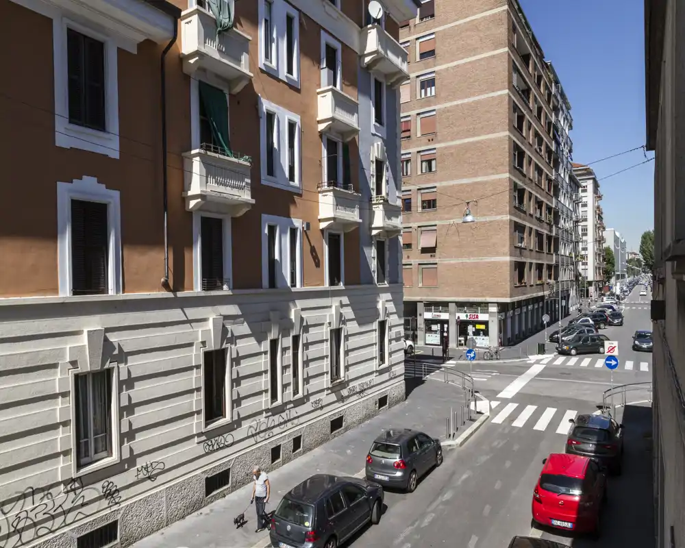 Veduta esterna verso la strada che mostra un incrocio urbano con auto parcheggiate, facciate di palazzi e passanti alla luce del giorno — utile per illustrare il contesto e il quartiere dell’edificio.