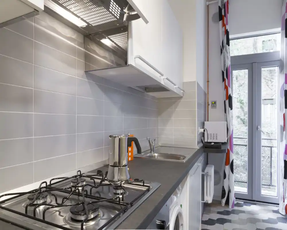 Cucina moderna e luminosa con piano cottura a gas a quattro fuochi, lavello in acciaio, piccoli elettrodomestici sul piano e porta sul balconcino — pulita, ben illuminata e funzionale per affitti brevi.