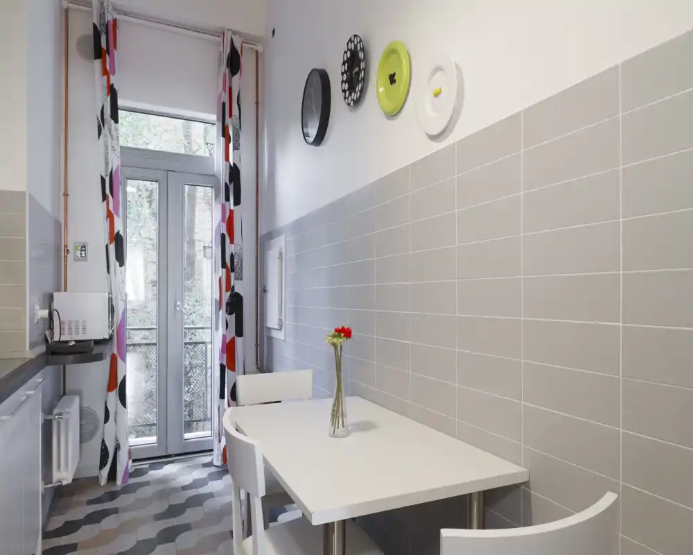 Angolo cucina compatto e moderno con un piccolo tavolo bianco, due sedie e tende decorative; luce naturale che entra dalla porta finestra verso il balcone.
