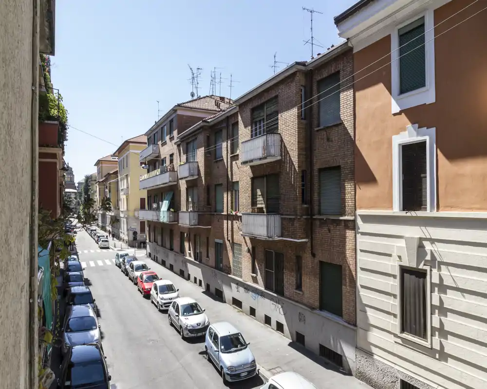 Vista sulla strada da un balcone/finestra che mostra una tranquilla via residenziale con auto parcheggiate e condomini di altezza media sotto un cielo terso.