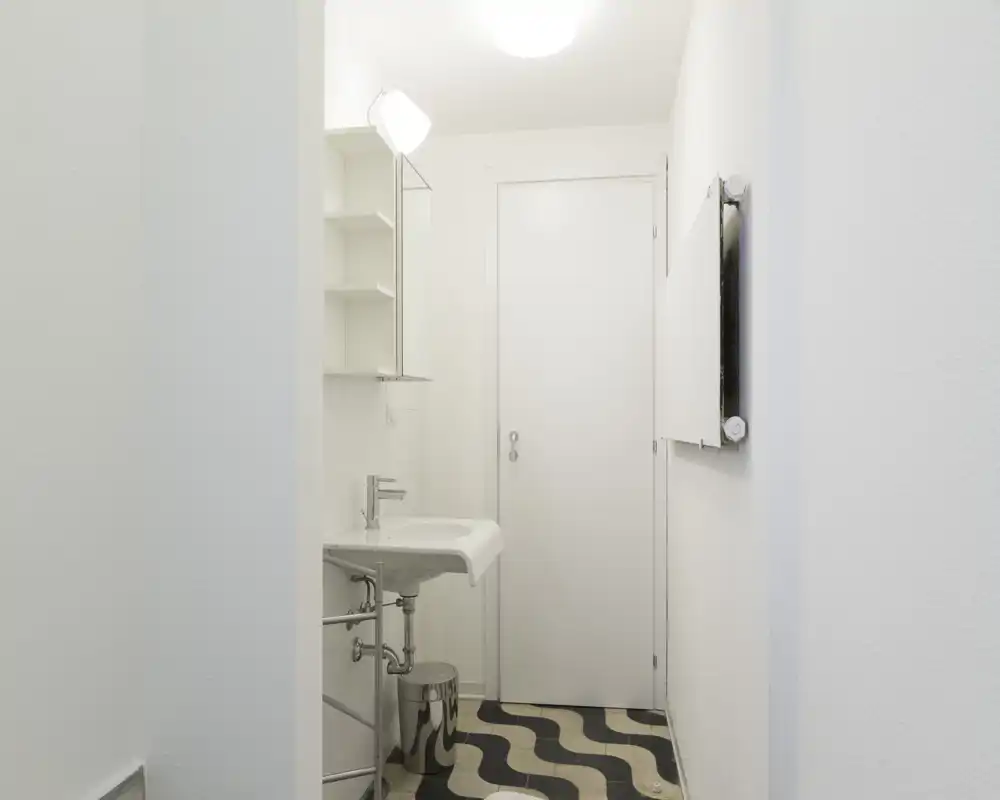 Bagno compatto e luminoso con lavabo a muro, mobile specchiera e pavimento piastrellato a motivo; stile pulito e minimal con buona illuminazione.