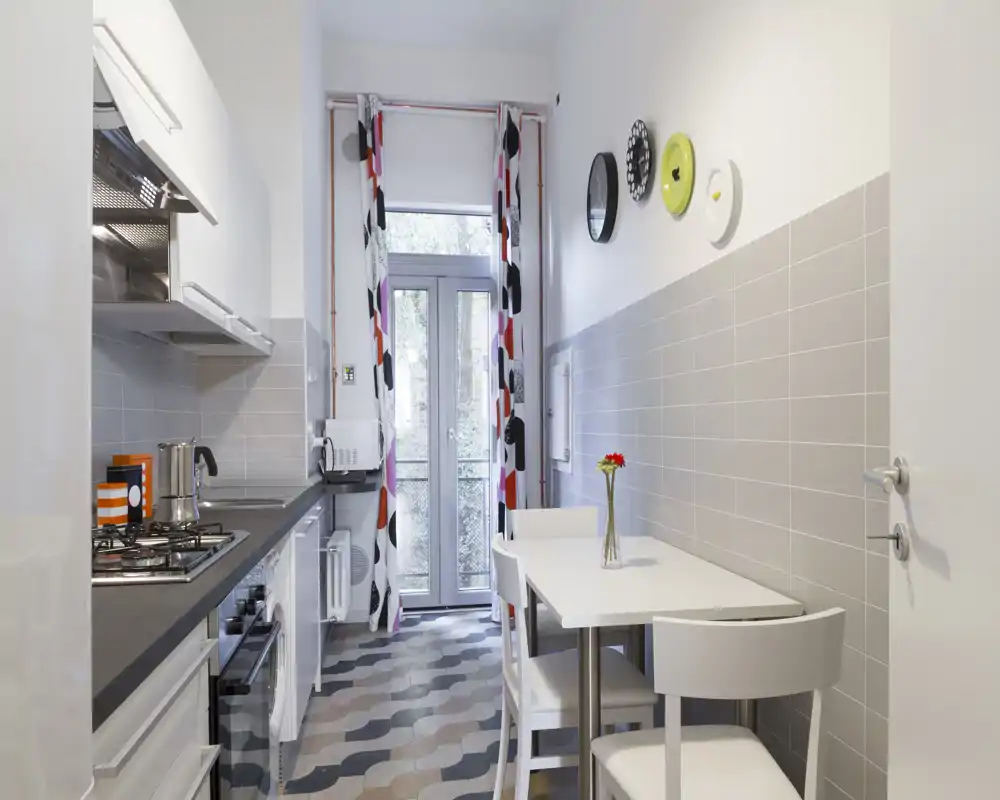 Cucina stretta e luminosa con tavolo da pranzo compatto, piano cottura integrato e mobili moderni. Tende colorate e dettagli a parete donano personalità, mentre la porta in vetro assicura luce naturale.