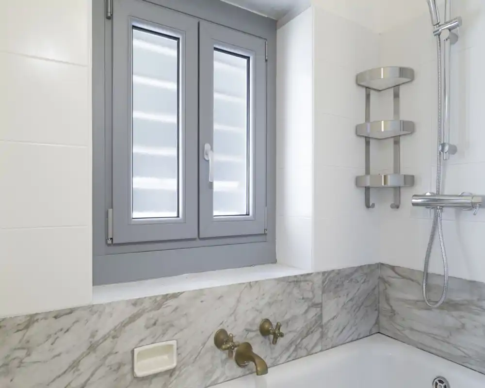 Angolo di bagno moderno e luminoso con vasca e rivestimento in marmo, rubinetteria in ottone, finestra a vetro satinato e colonna doccia cromata.