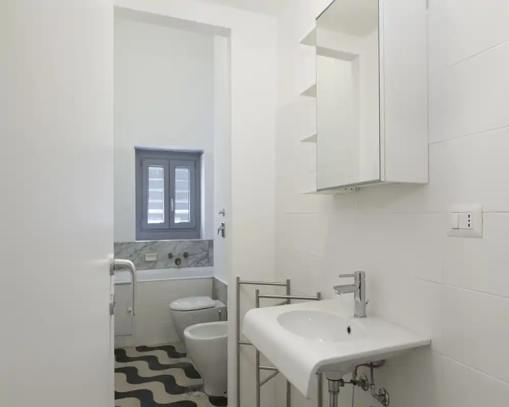 Bagno moderno e luminoso con lavabo sospeso, specchiera con pensile, bidet e wc. Piastrelle bianche e pavimento decorato creano un aspetto contemporaneo e pulito.