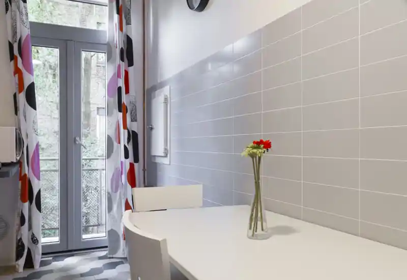 Bright, modern dining corner with a white table, simple chairs and a small vase of flowers beside a glass door leading to a balcony. Grey tiled wall and patterned curtains give a clean, contemporary look.
