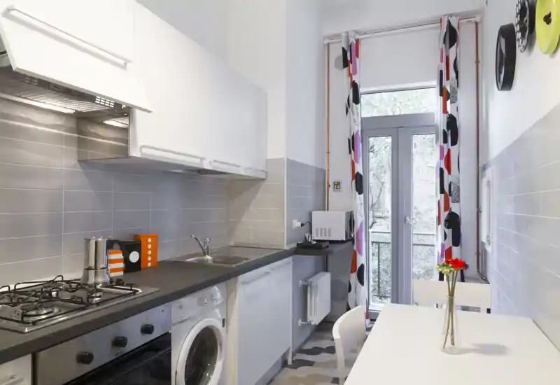 Modern narrow kitchen with built-in stove, sink, washing machine and a small dining table by a glass door letting in natural light.