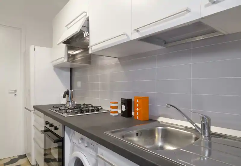 Compact, modern kitchen featuring a stainless sink, gas hob, built-in oven and refrigerator. Neutral grey tiles and white cabinets give a clean, contemporary look.