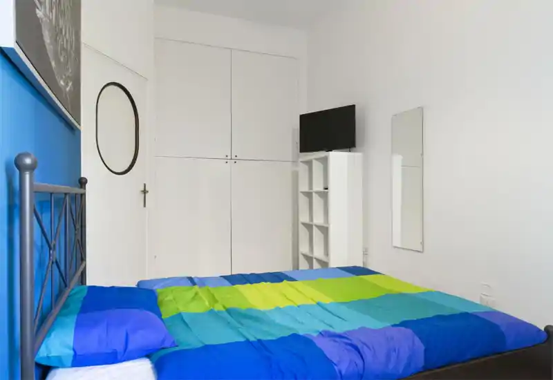 Chambre lumineuse et compacte avec un lit double à cadre métallique et une couette bleu-vert, une armoire encastrée et une petite étagère avec TV — propre et prête pour les hôtes.