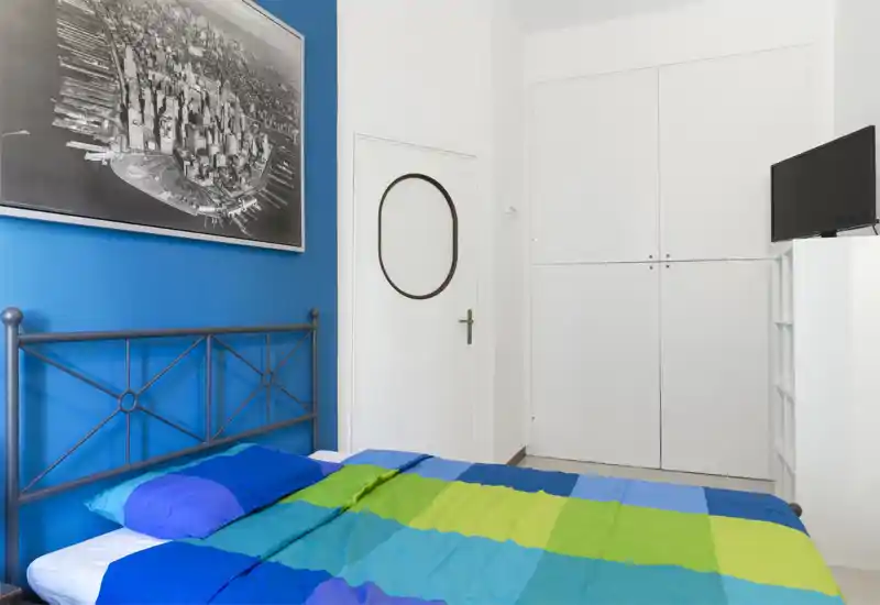 Chambre lumineuse et bien rangée avec tête de lit en métal, linge de lit coloré et grande affiche encadrée sur un mur bleu. Rangements encastrés et petite télévision pour un séjour pratique.