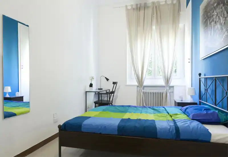 Chambre lumineuse et sobre avec un lit double à la literie colorée, une table de chevet avec lampe, un petit bureau et une chaise près de la fenêtre, apportant beaucoup de lumière naturelle.