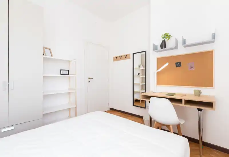 Camera da letto luminosa e minimal con letto rifatto in primo piano, una postazione di lavoro compatta con scrivania e sedia a destra e armadio con scaffali a sinistra.