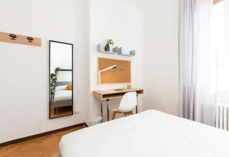 Camera da letto luminosa e minimal con letto ben rifatto, una scrivania in legno e uno specchio a figura intera. La luce naturale e l’arredo ordinato creano un’atmosfera tranquilla e accogliente.