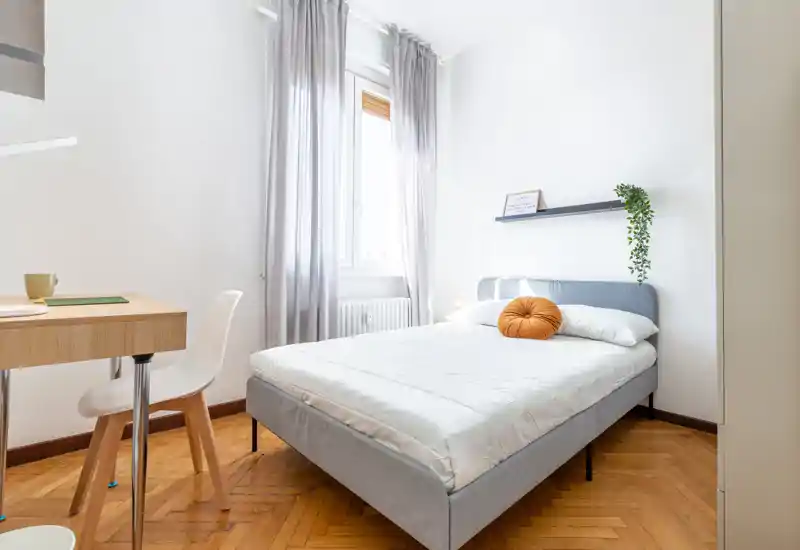 Camera da letto moderna e luminosa con letto ben rifatto, testiera grigia, cuscino decorativo rotondo e una scrivania minimal vicino alla finestra. La luce naturale e il pavimento in legno creano un ambiente accogliente, perfetto per annunci di affitto.