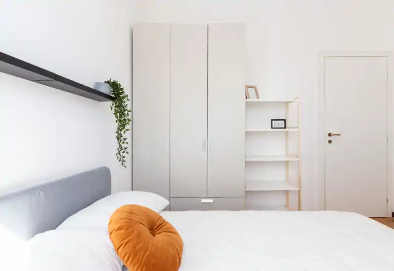 Camera da letto luminosa e minimal: letto ben fatto con testiera grigio chiaro, biancheria bianca e cuscino arancione decorativo; scaffalatura in legno chiaro e armadio che donano un aspetto pulito e arioso.