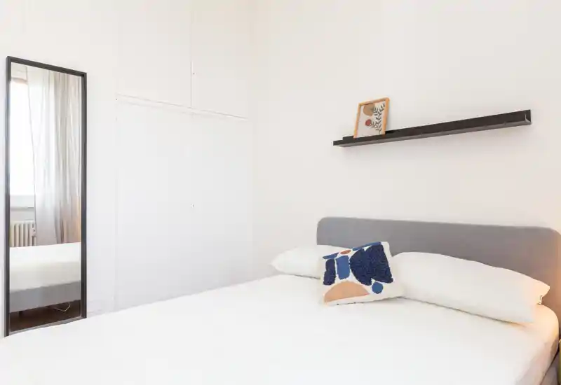 Camera da letto luminosa e minimalista con letto matrimoniale, biancheria neutra e un cuscino decorativo; uno specchio alto e una mensola completano lo spazio per un aspetto moderno e pulito.