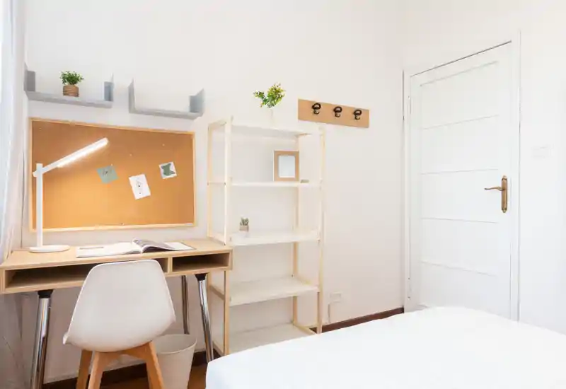 Angolo camera luminoso e minimal con scrivania, scaffale e parte del letto. Pareti bianche e luce naturale creano un ambiente ordinato e funzionale, ideale per studiare e riposare.