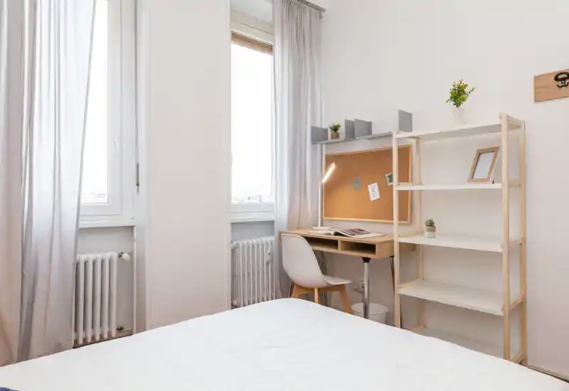 Camera da letto luminosa e minimal con letto ben rifatto in primo piano e un angolo studio compatto con scrivania e scaffale vicino alla finestra.