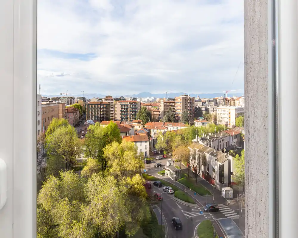Vista cittadina dalla finestra con strade alberate, edifici residenziali e colline lontane sotto un cielo luminoso.