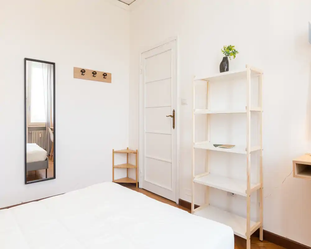 Camera da letto minimale e luminosa con letto bianco ben rifatto, mensole in legno e uno specchio a figura intera che riflette parte della stanza. Ambiente tranquillo e ben illuminato ideale per annunci.